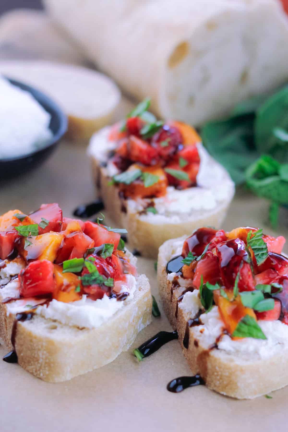 Three assembled strawberry and peach bruschetta toast drizzled with honey and balsamic glaze. There is a bowl of goat cheese to the top left of the bruschetta with the baguette at the top and fresh min and basil to the top right of the bruschetta.