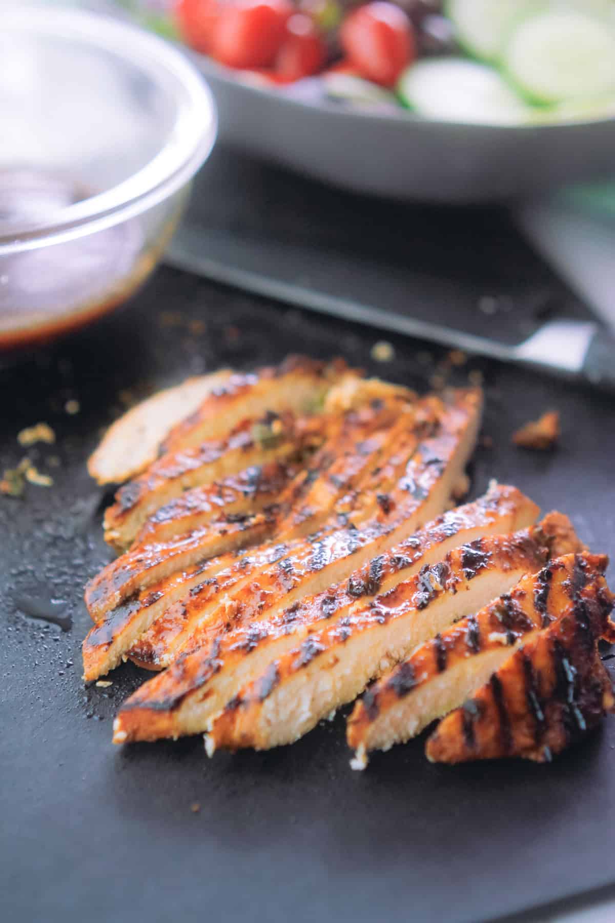 Sliced grilled chicken breast with a bowl of fresh vegetables at the top right corner of sliced chicken.