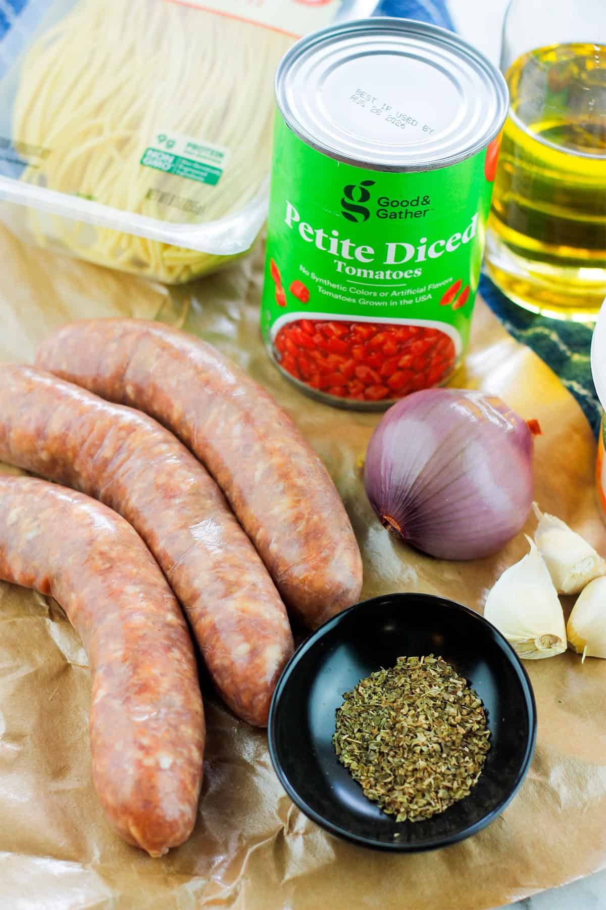 Ingredients for sausage pasta clockwise from the top a can do petite diced tomatoes, glass jar of olive oil, fresh shallot and garlic cloves, small black bowl filled with italian herbs, three links of sweet italian sausage, and a package of fresh uncooked spaghetti pasta.