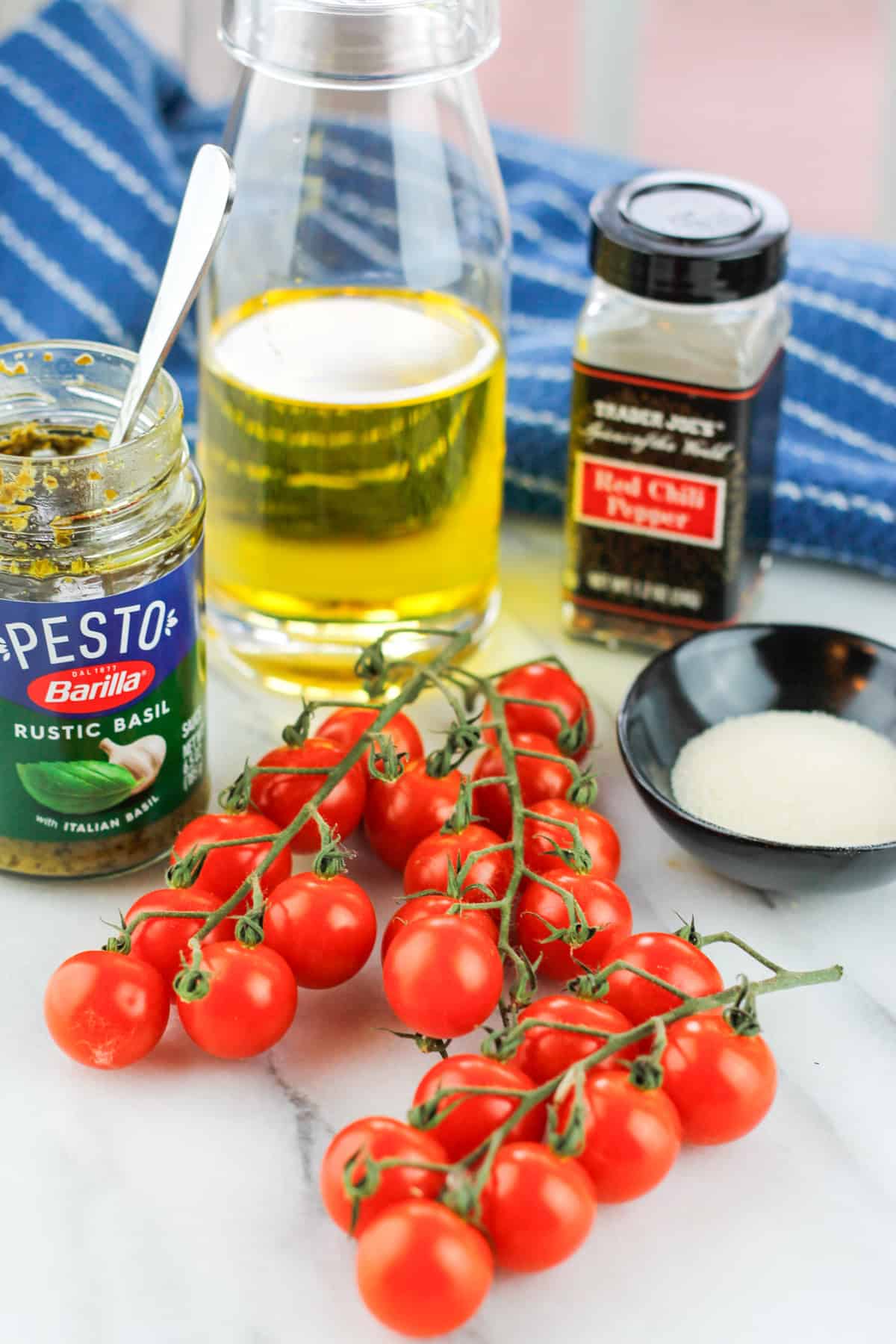 Counterclockwise shot of ingredients cherry tomatoes in the middle jar of pesto sauce with a spoon inserted in jar glass jar of olive oil jar of red pepper flakes and small black bowl of sugar all with a blue towel in the background.