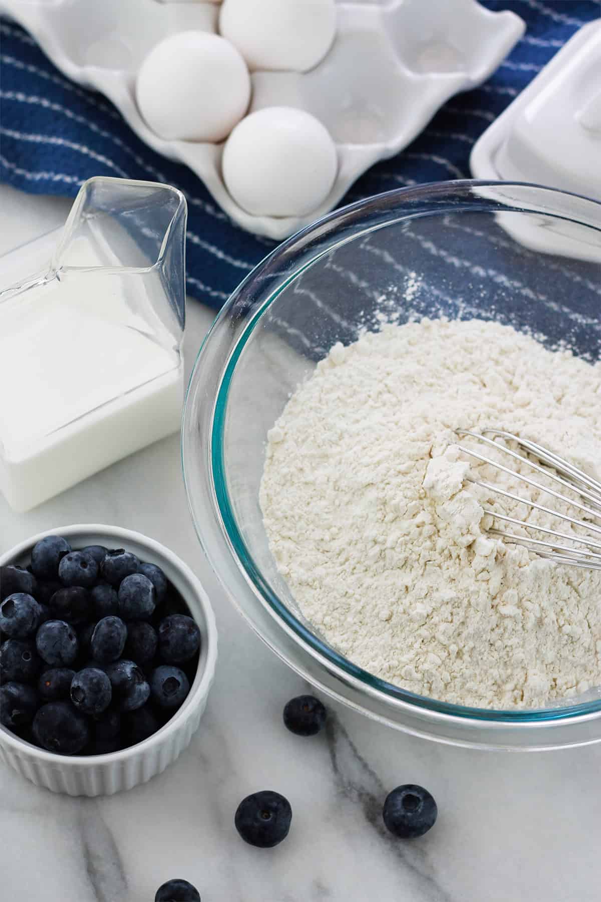 Overhead shot going counterclockwise of small bowl of fresh blueberries flour in a glass mixing bowl butter dish egg crate and glass container of milk on a marble counter.