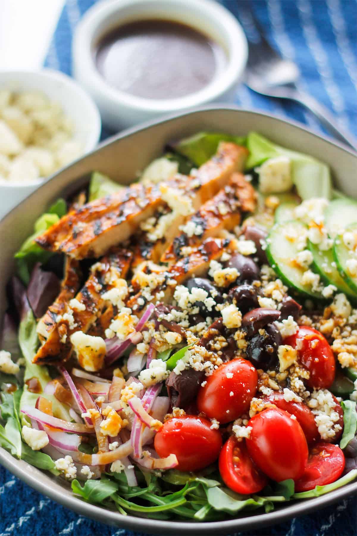 Sliced grilled chicken atop a salad with sliced cucumbers, sliced grape tomatoes, olives, sliced red onions, on a bed of mixed greens, garnished with crumbled cheese.