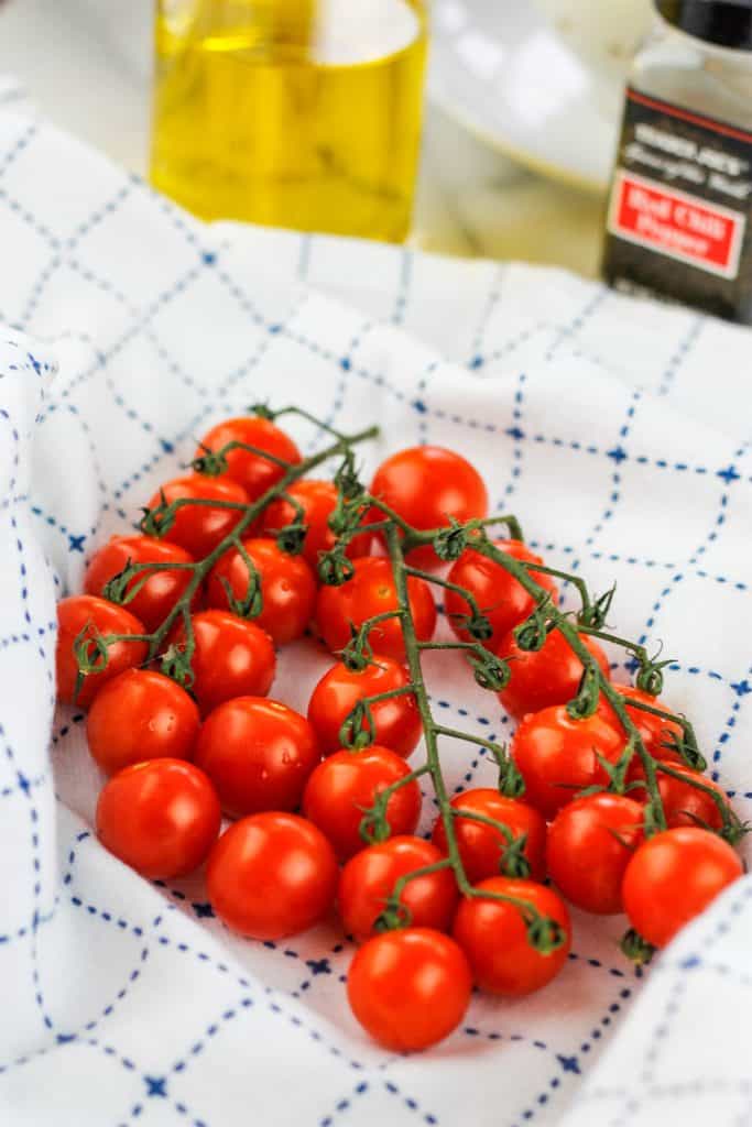 Washed cherry tomatoes on a blue and white towel with glass jar of olive oil and jar of red pepper flakes in the background