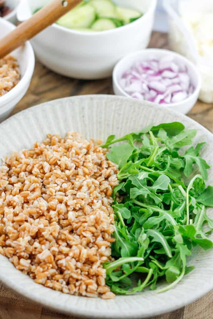 Bowl filled with cooked farro on the left and fresh arugula on the right with bowls of chopped vegetables behind the grain bowl.