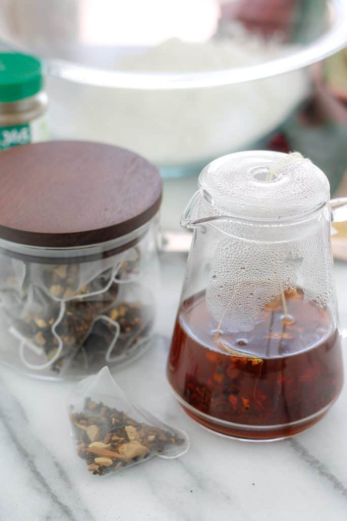 Chai tea steeping in small glass pitcher on the right and a glass container with chai tea sachets on the left with a glass mixing bowl with dry ingredients in the background