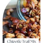 Glass container of chipotle trail mix on its side with the trail mix spilling out onto the wood table.