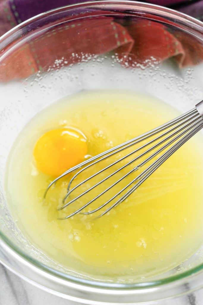 Glass bowl with egg being mixed into sugar mixture with a wire whisk with a dish towel behind the bowl.