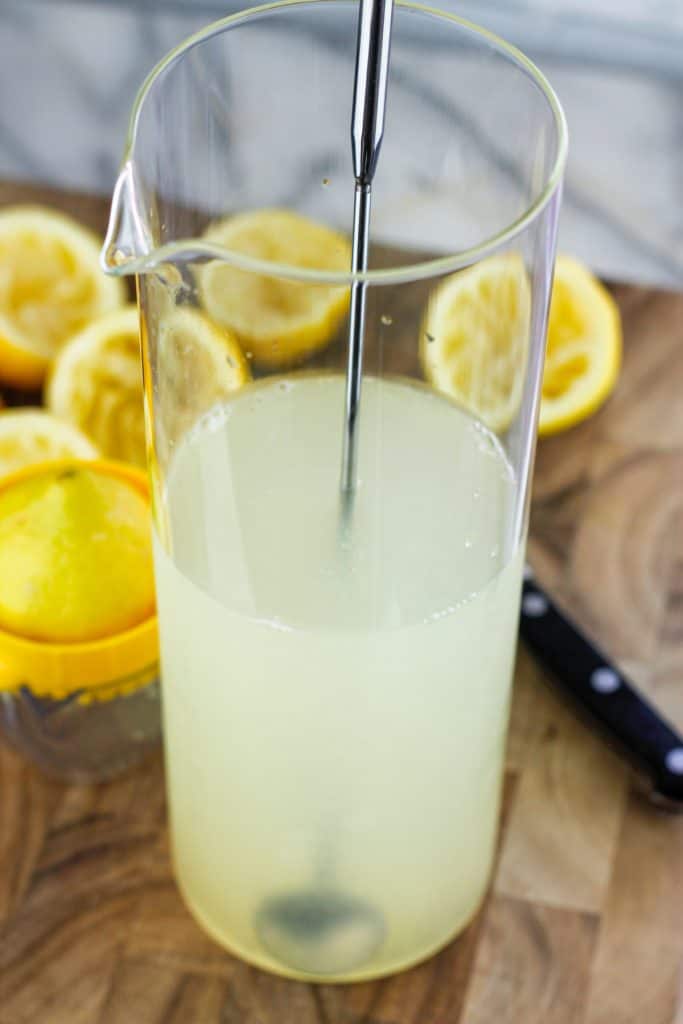 Glass pitcher filled with fresh water and lemon juice with mixing spoon in pitcher. Sliced and squeezed lemons around the pitcher.