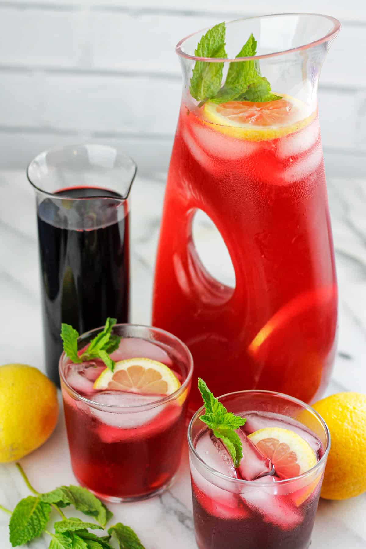 Glass pitcher of hibiscus lemonade and two short glasses of hibiscus lemonade in front of pitcher with a glass container of hibiscus syrup next to large container of hibiscus lemonade.