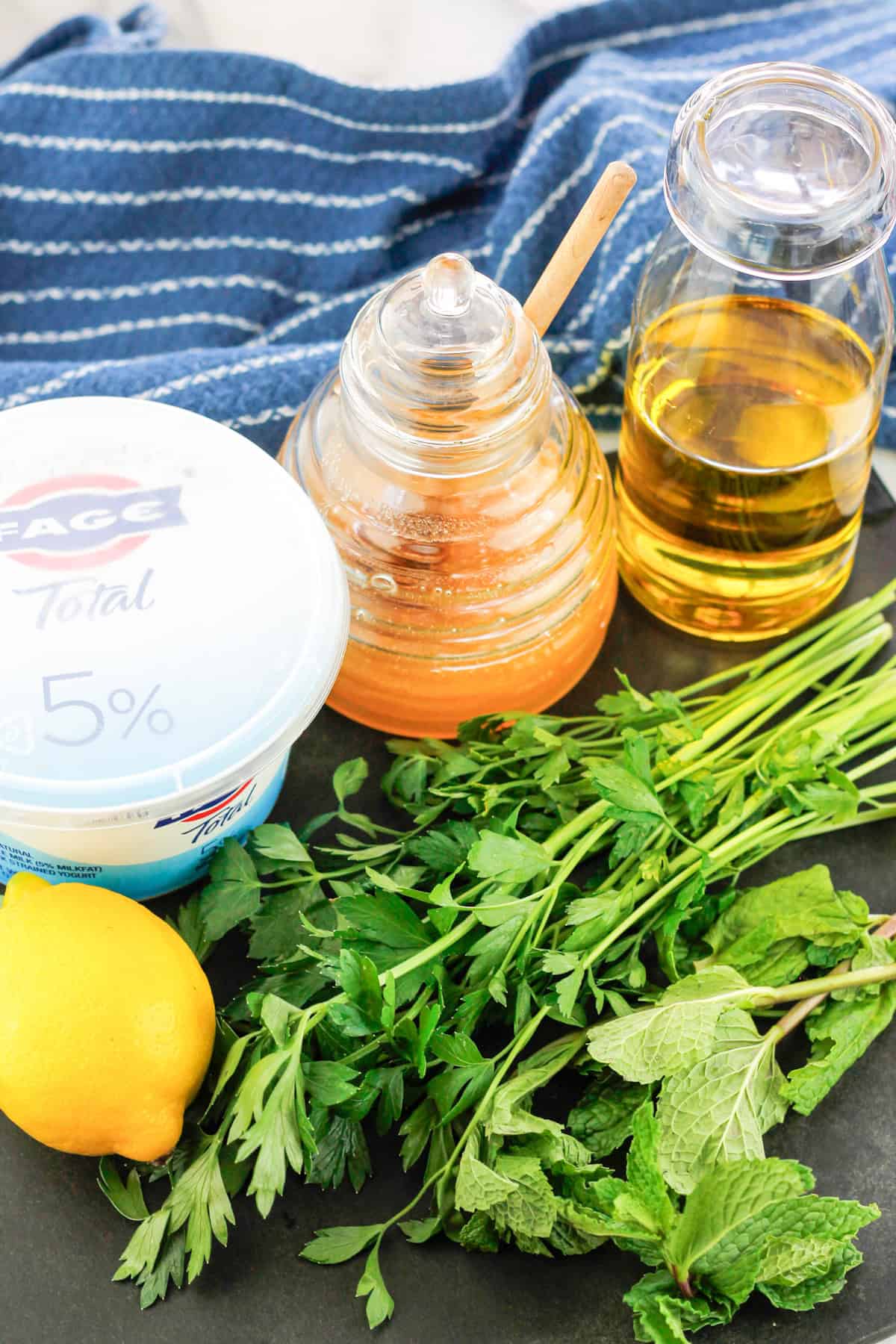 Mint and flat leaf parsley bottom right with a lemon to the left and a container of greek yogurt behind the lemon with a honey jar next to the yogurt container and a glass container of olive oil next to the honey jar.