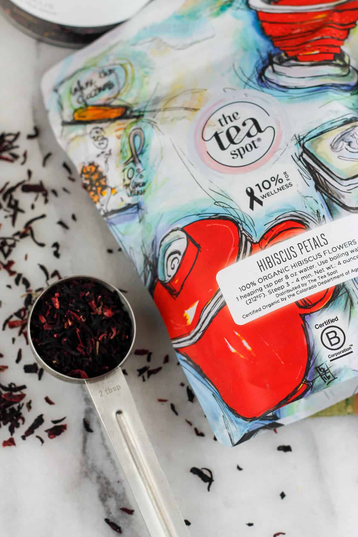 Overhead shot of bag of loose leaf hibiscus tea flowers next to a scoop of hibiscus tea flowers on a marble countertop