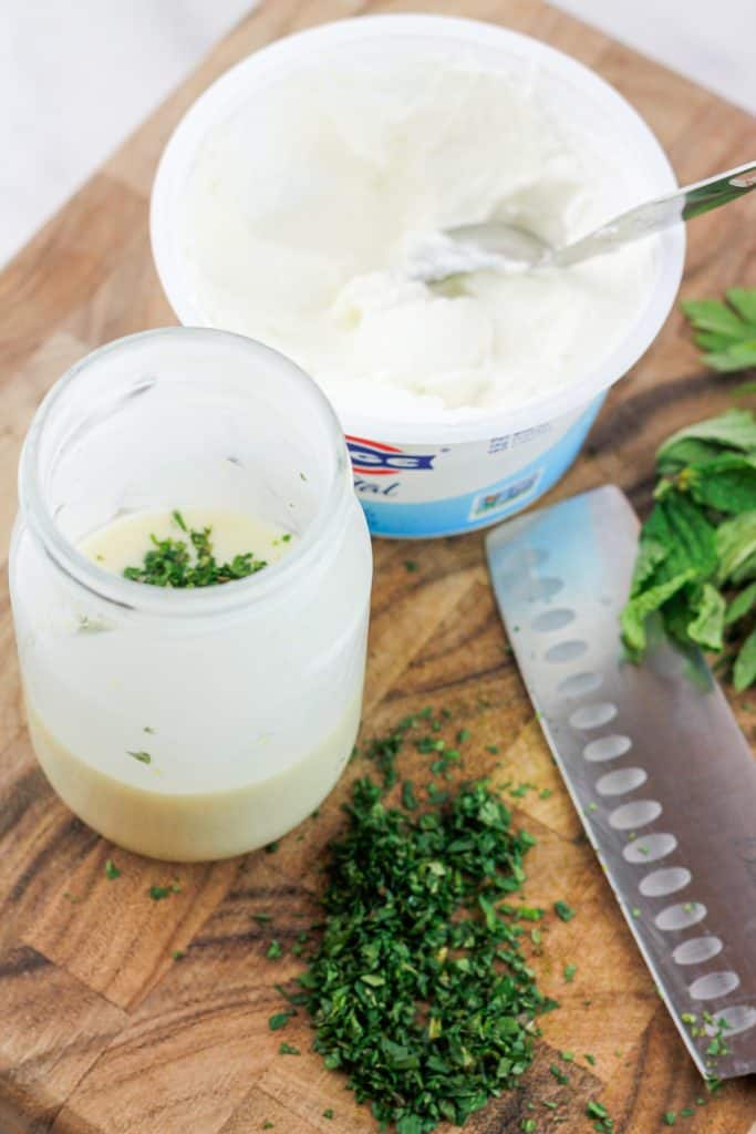 Overhead shot of dressing in a jar with chopped herbs and a container of greek yogurt on a wood cutting board.