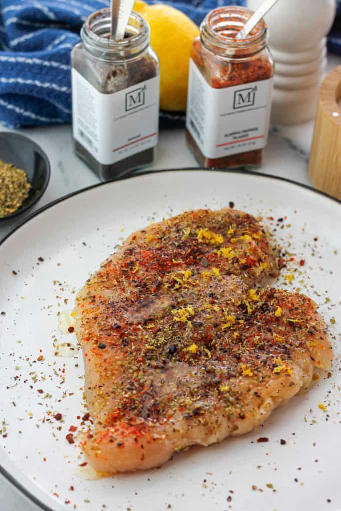 Seasoned chicken breast on a white plate with jared seasoning behind the plate.