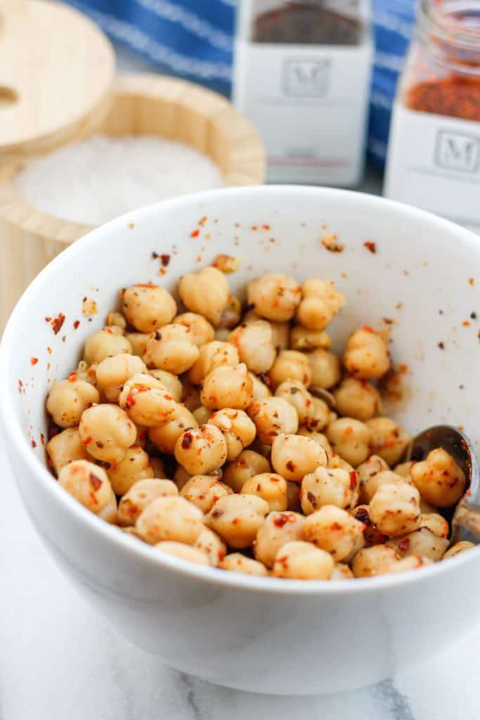 Seasoned chickpeas in a bowl with a spoon a salt cellar and jars of spices behind the bowl.