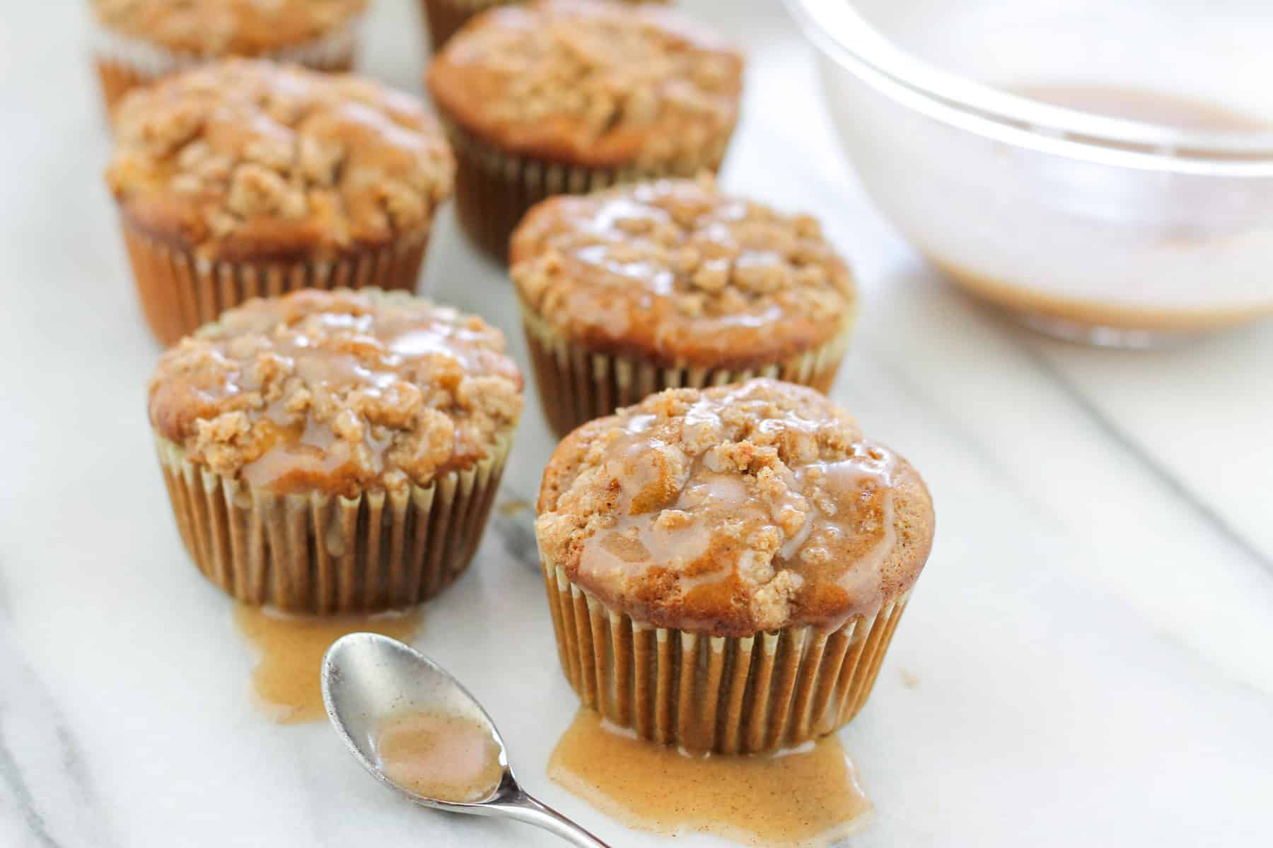 Shot of chai apple muffin on counter with the front muffins topped with some of the chai glaze with a small spoon filled with chai sugar glaze.