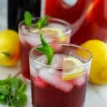 Two short glasses filled with hibiscus lemonade with hibiscus syrup and large container of hibiscus behind the short glasses.