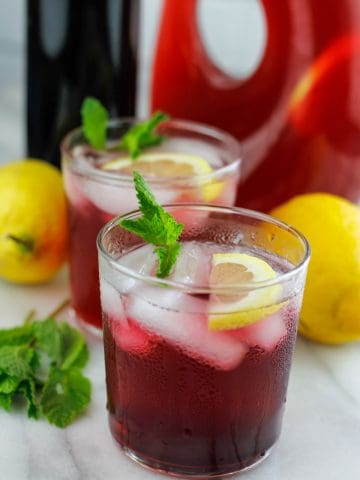 Two short glasses filled with hibiscus lemonade with hibiscus syrup and large container of hibiscus behind the short glasses.