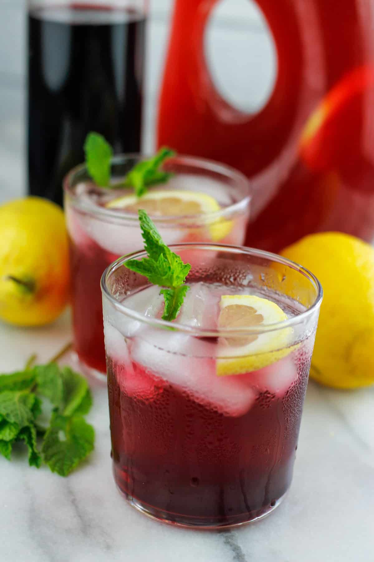 Two short glasses filled with hibiscus lemonade with hibiscus syrup and large container of hibiscus behind the short glasses.