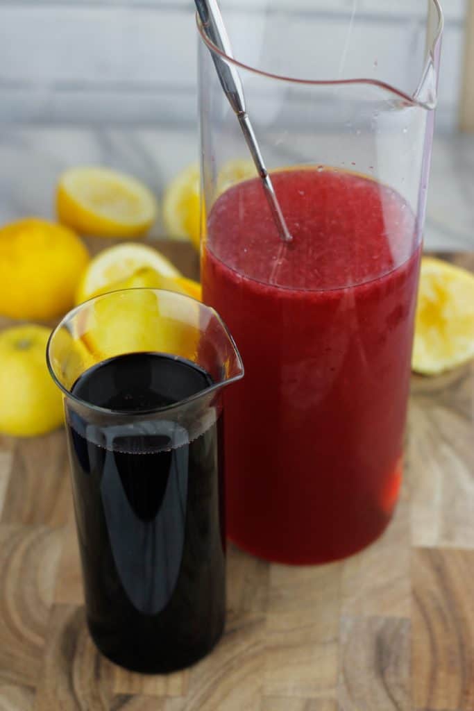 small glass container filled with hibiscus syrup on the left and a larger glass container of hibiscus lemonade on the right.