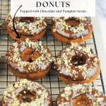 Pumpkin donuts topped in chocolate and chopped pumpkin seeds on a cooling rack.