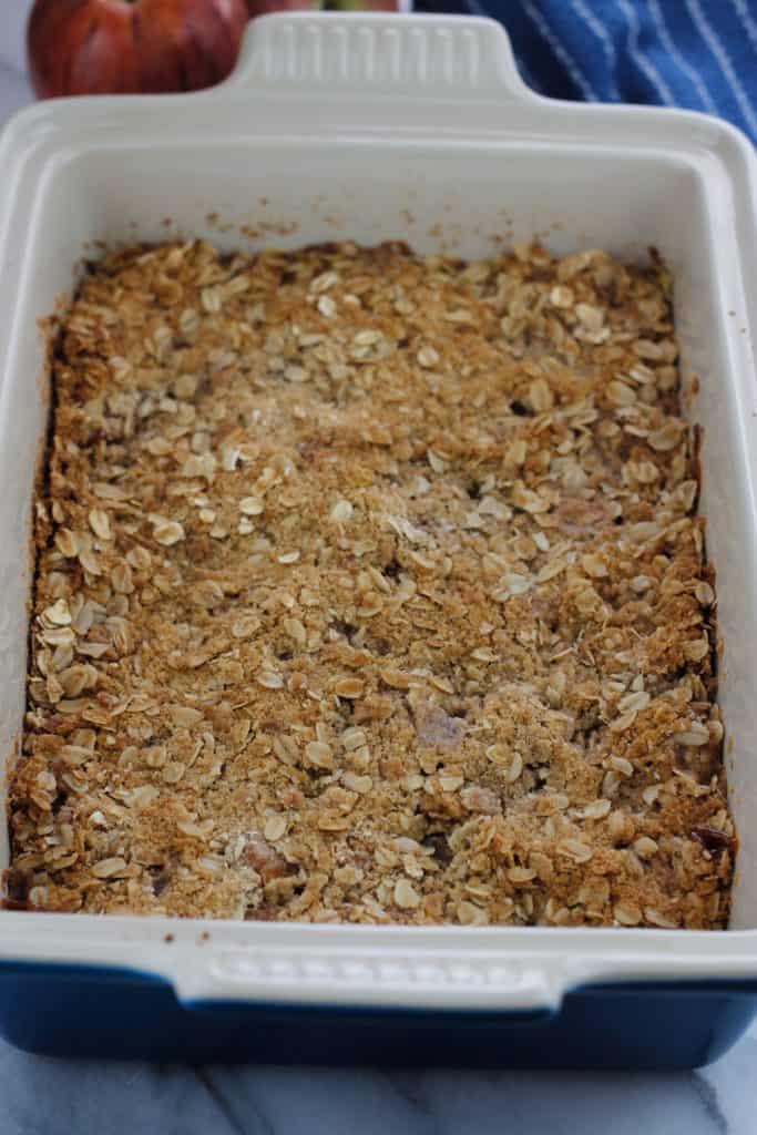 Baked apple crisp in a rectangle baking dish with two fresh apples at the top left of the baking dish.