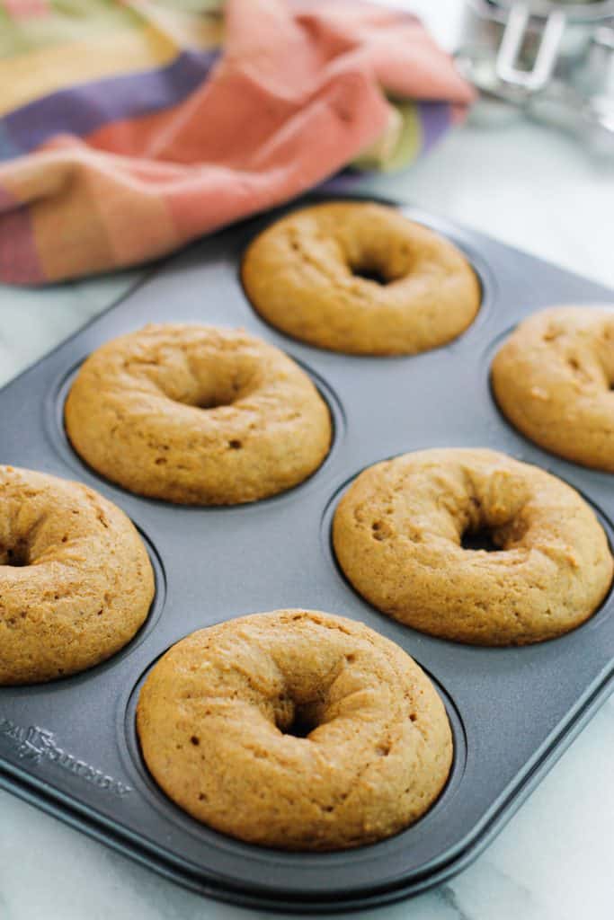 Baked pumpkin donut cooling in the donut pan on a marble countertop with a dish towel to the left of the pan.