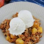 Bowl of pumpkin apple crisp with two scoops of ice cream there are two fresh apples at the back left of the bowl and the corner of the baking dish at the back right of the bowl.