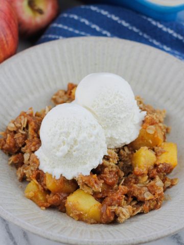 Bowl of pumpkin apple crisp with two scoops of ice cream there are two fresh apples at the back left of the bowl and the corner of the baking dish at the back right of the bowl.