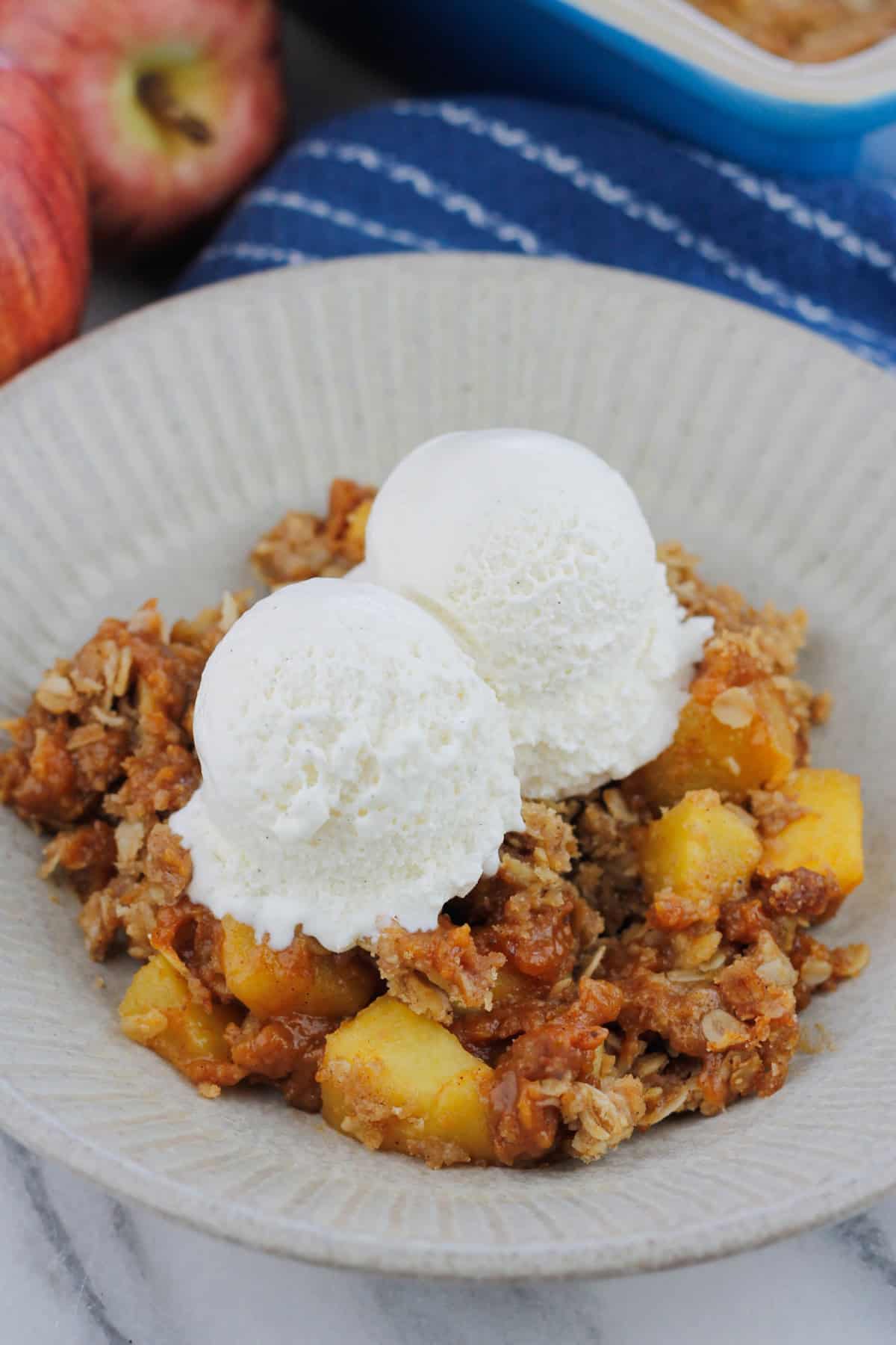 Bowl of pumpkin apple crisp with two scoops of ice cream there are two fresh apples at the back left of the bowl and the corner of the baking dish at the back right of the bowl.