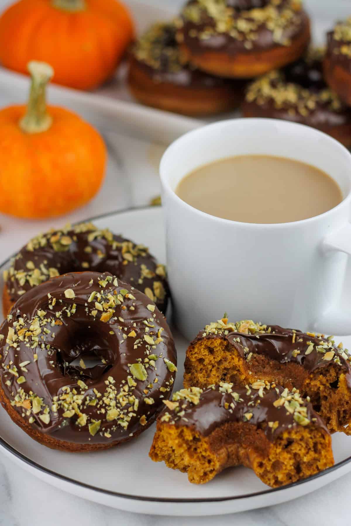 Chocolate dipped pumpkin donuts sprinkled with chopped pumpkin seeds on a plate with a cup of coffee.