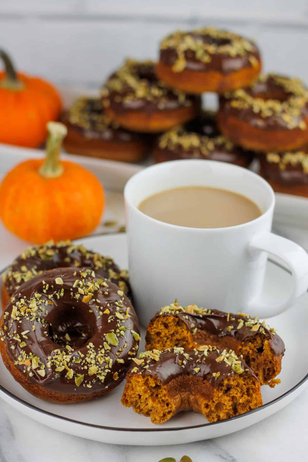 Chocolate dipped pumpkin donuts sprinkled with chopped pumpkin seeds on a plate with a cup of coffee on a plate.