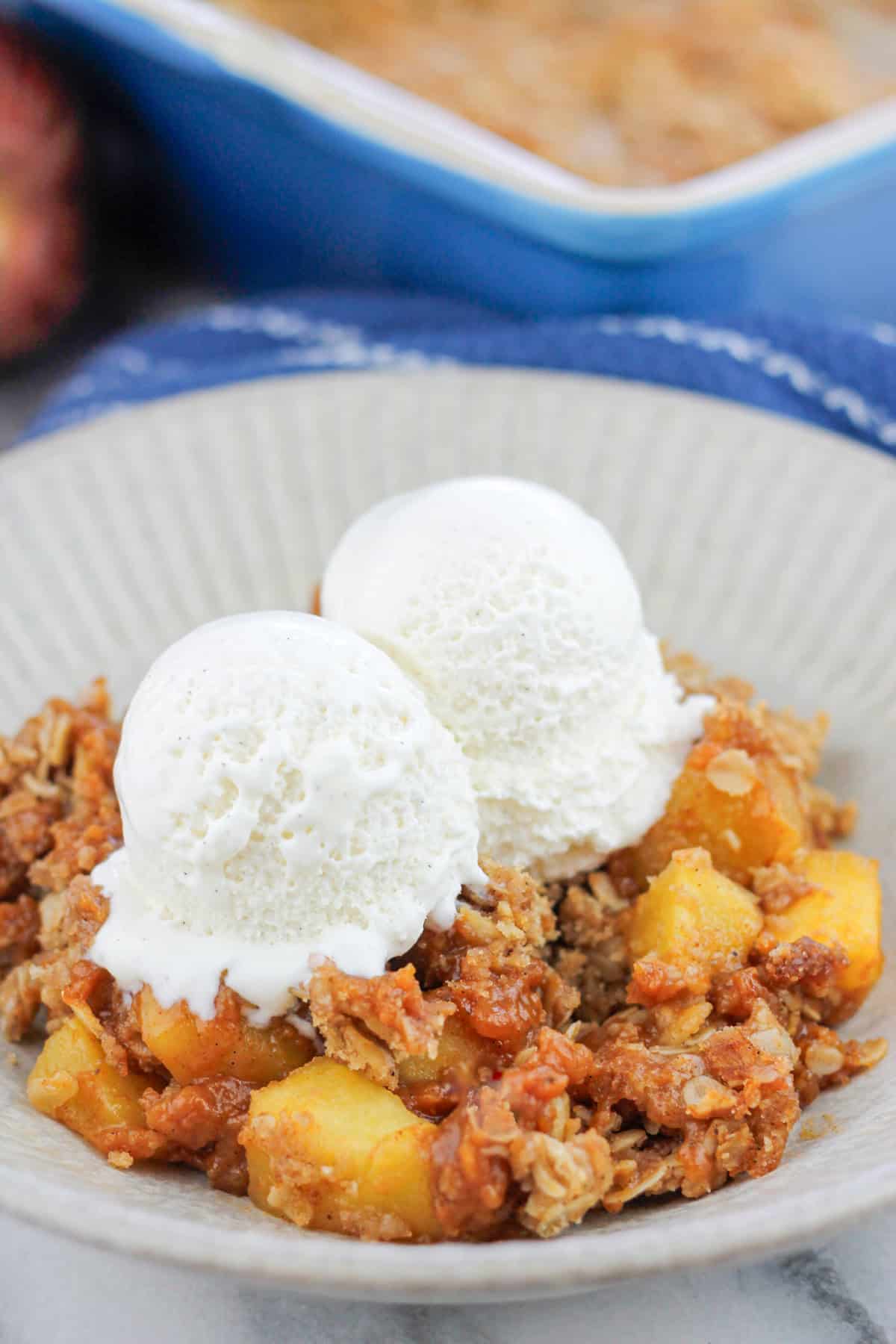 Close-up shot of a bowl of pumpkin apple crisp with two scoops of ice cream there are two fresh apples at the back left of the bowl and the corner of the baking dish at the back right of the bowl.