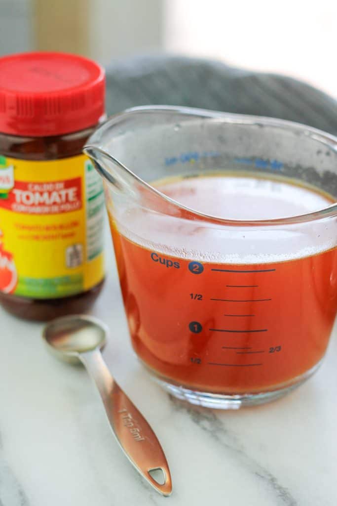 Glass measuring cup filled with tomato stock with a container of Knorr tomato bouillon to the left of the measuring cup.