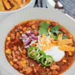 Overhead shot of easy rotisserie chicken tortilla soup in a bowl, the soup is garnished with sour cream, diced red onions, shredded cheese, three jalapeño slices, cilantro, and fried tortilla strips. Stems of fresh cilantro leaves are on the table at the bottom right of the bowl and a bowl of fried tortilla strips are at the top left of the bowl. A silver spoon is at the top right of the bowl on a grey striped napkin.