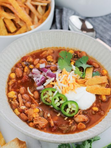 Overhead shot of easy rotisserie chicken tortilla soup in a bowl, the soup is garnished with sour cream, diced red onions, shredded cheese, three jalapeño slices, cilantro, and fried tortilla strips. Stems of fresh cilantro leaves are on the table at the bottom right of the bowl and a bowl of fried tortilla strips are at the top left of the bowl. A silver spoon is at the top right of the bowl on a grey striped napkin.