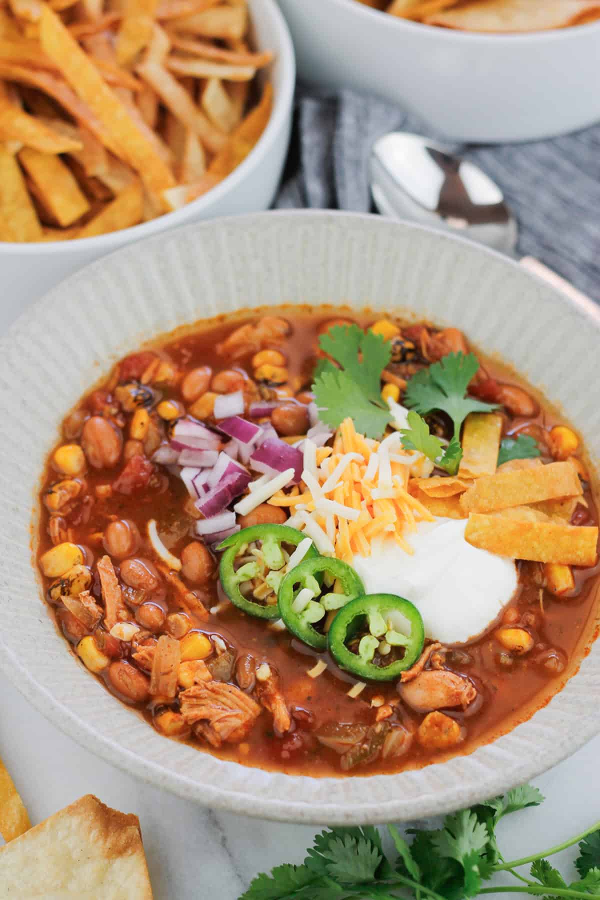 Overhead shot of easy rotisserie chicken tortilla soup in a bowl, the soup is garnished with sour cream, diced red onions, shredded cheese, three jalapeño slices, cilantro, and fried tortilla strips. Stems of fresh cilantro leaves are on the table at the bottom right of the bowl and a bowl of fried tortilla strips are at the top left of the bowl. A silver spoon is at the top right of the bowl on a grey striped napkin.