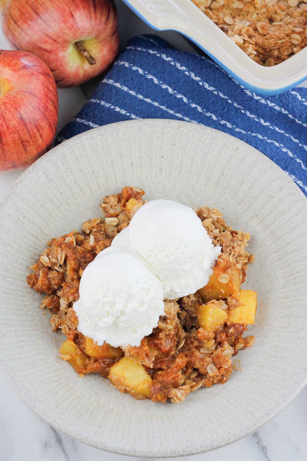 Overhead shot of a bowl of pumpkin apple crisp with two scoops of ice cream there are two fresh apples at the back left of the bowl and at the top right of the bowl the corner of the baking dish with the baked pumpkin apple crisp.