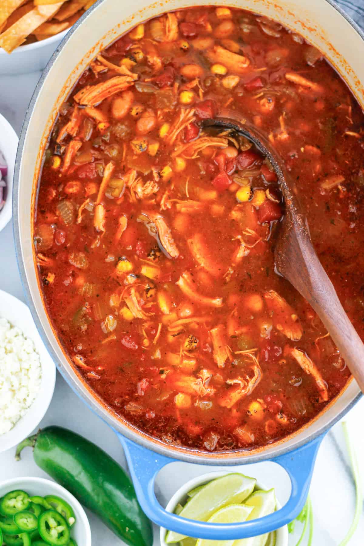 Overhead shot of tortilla soup in pot with wooden ladle in soup and bowls of chopped vegetables and cheese for garnishing around the left side of the pot.