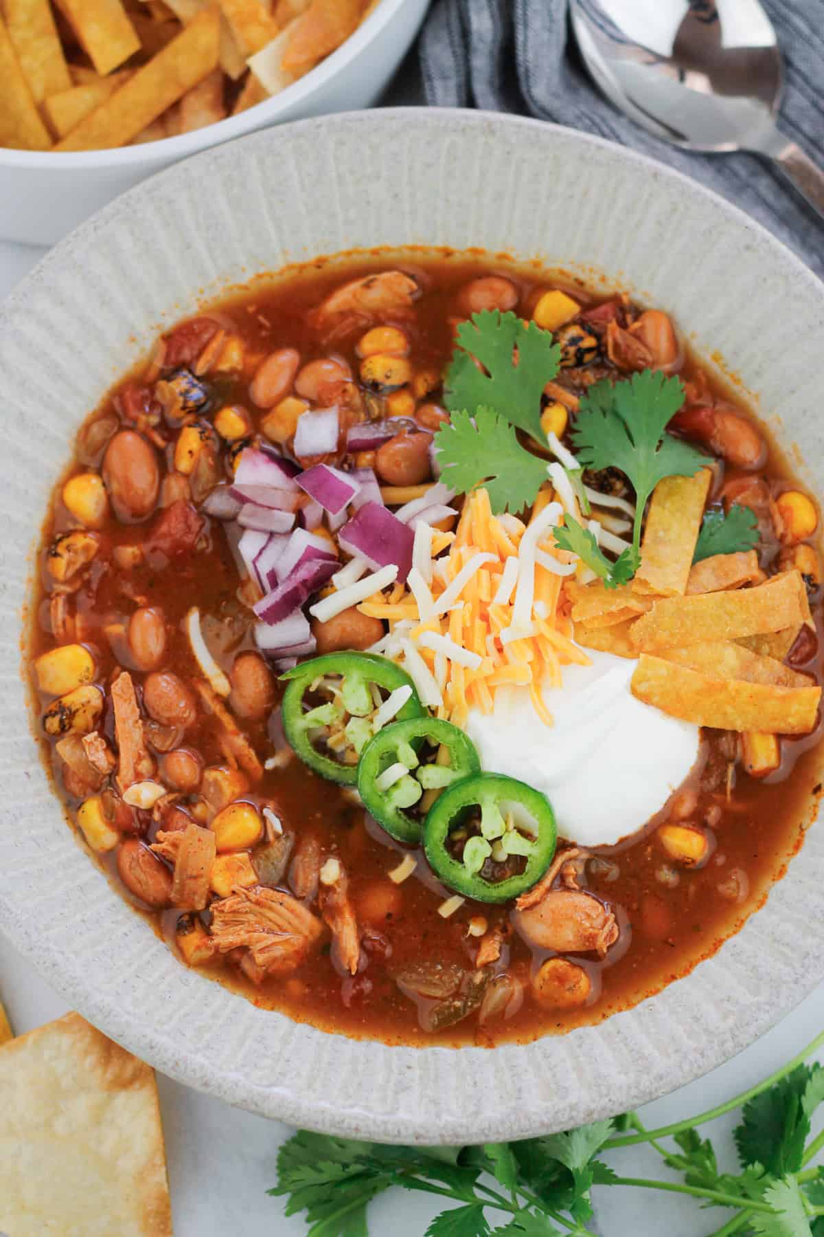 Overhead shot of easy rotisserie chicken tortilla soup in a bowl, the soup is garnished with sour cream, diced red onions, shredded cheese, three jalapeño slices, cilantro, and fried tortilla strips. Stems of fresh cilantro leaves are on the table at the bottom right of the bowl and a bowl of fried tortilla strips are at the top left of the bowl. A silver spoon is at the top right of the bowl on a grey striped napkin.