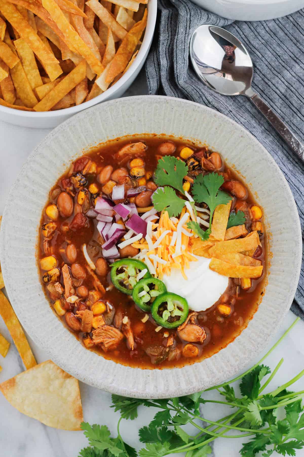 Overhead shot of easy rotisserie chicken tortilla soup in a bowl, the soup is garnished with sour cream, diced red onions, shredded cheese, three jalapeño slices, cilantro, and fried tortilla strips. Stems of fresh cilantro leaves are on the table at the bottom right of the bowl and a bowl of fried tortilla strips are at the top left of the bowl. A silver spoon is at the top right of the bowl on a grey striped napkin.