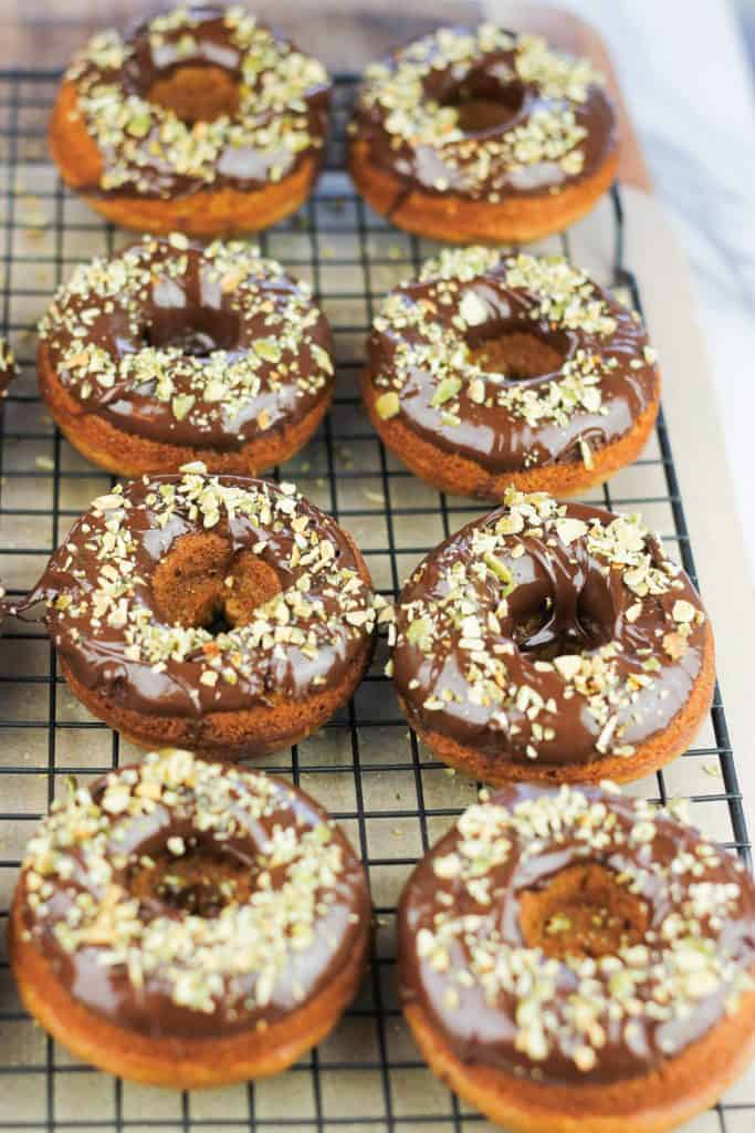 Pumpkin donuts topped in chocolate and chopped pumpkin seeds on a cooling rack.