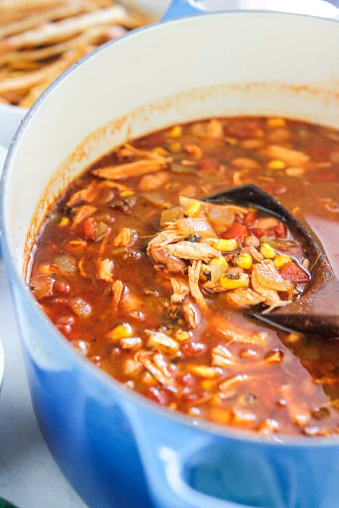 Shot of rotisserie chicken tortilla soup in dutch oven with a wooden ladle in soup and crispy tortilla strip to the left back of the pot.