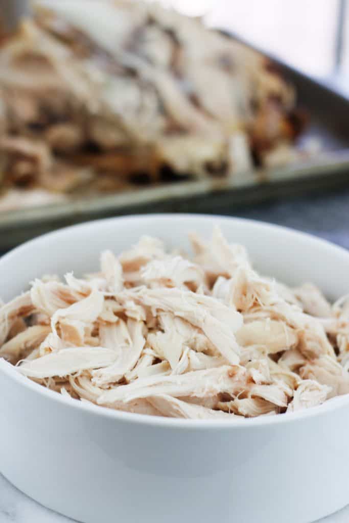 Shredded chicken in a white bowl from a rotisserie cooked chicken with the bones of the chicken on a pan in the background.