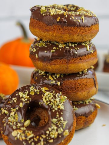 Stacked chocolate topped pumpkin donuts stacked on a plate with one donut propped on the stacked donuts.