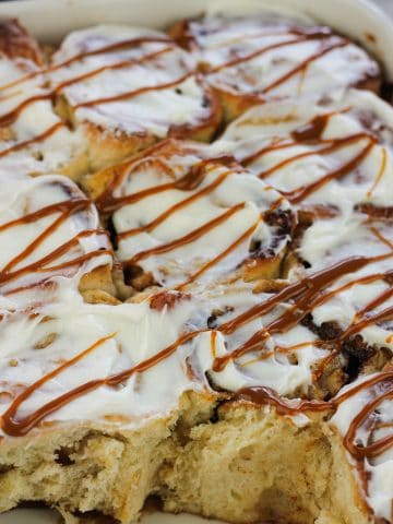 Shot of caramel apple cinnamon roll in a baking dish topped with cream cheese glaze and drizzled wth caramel sauce.