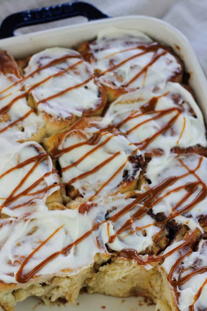 Close shot of caramel apple cinnamon roll in a baking dish topped with cream cheese glaze and drizzled wth caramel sauce.