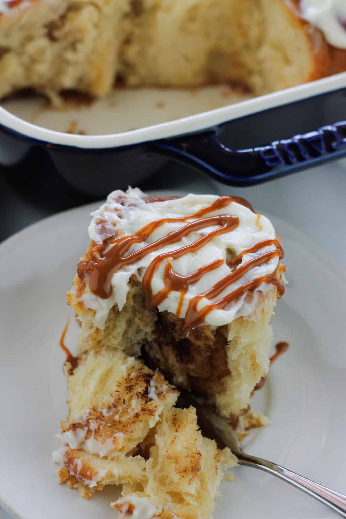 A single caramel apple cinnamon roll on a dish with cream cheese topping and a drizzle of caramel sauce on top.