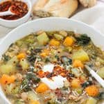 Overhead shot of a bowl of butternut white bean soup with Swiss chard and a small bowl of crispy chili onion oil and crusty bread of in the background.