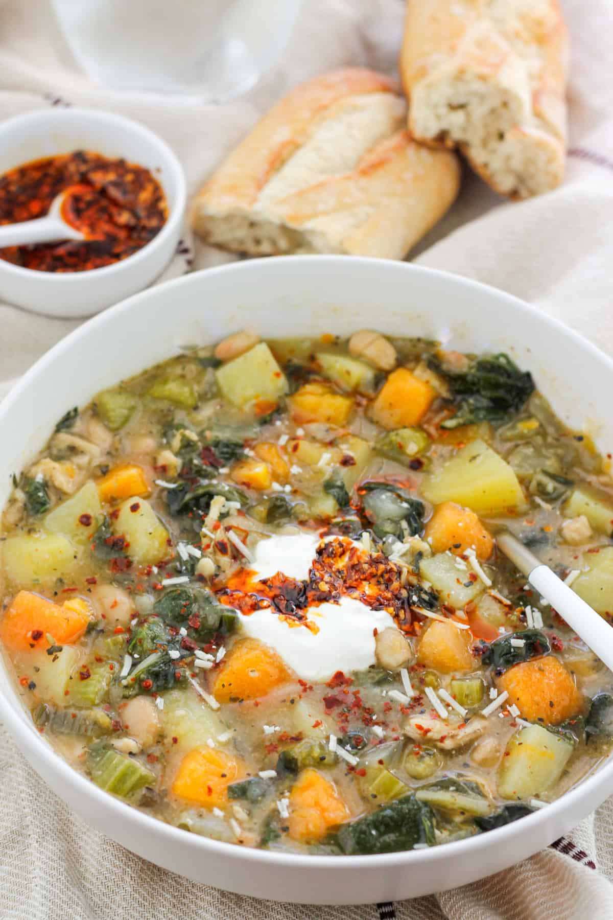 Overhead shot of a bowl of butternut white bean soup with Swiss chard and a small bowl of crispy chili onion oil and crusty bread of in the background.