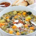 Close shot of a bowl of butternut white bean soup with Swiss chard and a small bowl of crispy chili onion oil and crusty bread of in the background.